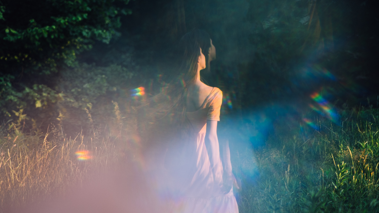 A woman standing peacefully in a sunlit grassy field surrounded by trees, with soft light prism reflections.